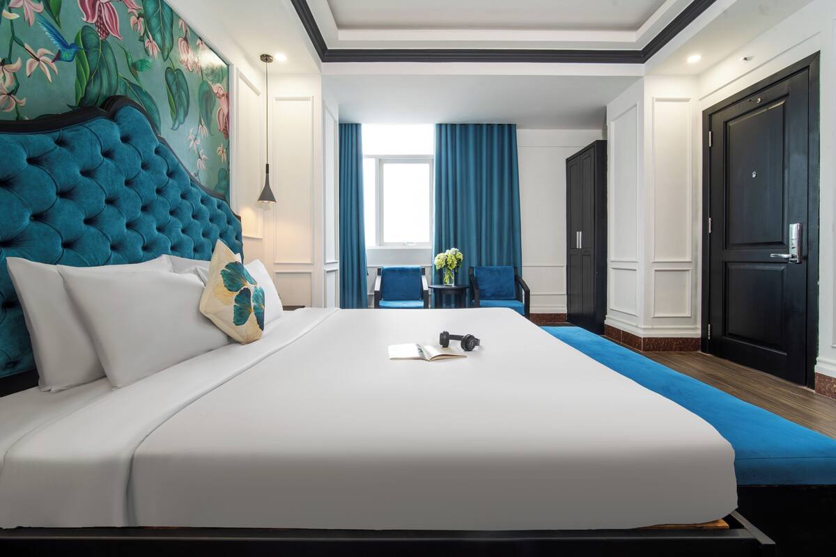 Habitación de hotel ordenada con una cama grande, un cabecero tapizado en azul, una mesita con un jarrón de flores y vistas al pasillo.