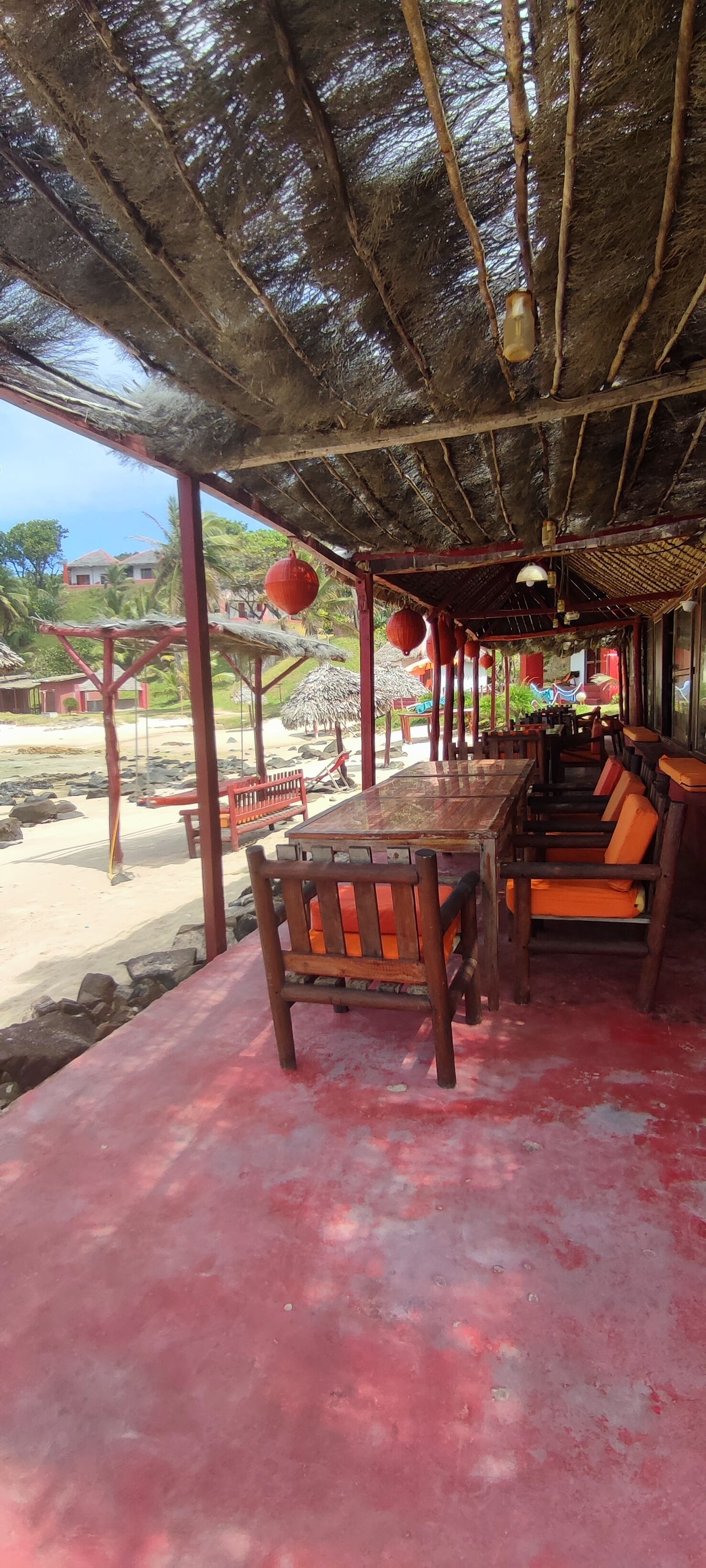 Un área de comedor al aire libre con techo de paja, mesas de madera y sillas con cojines naranja.