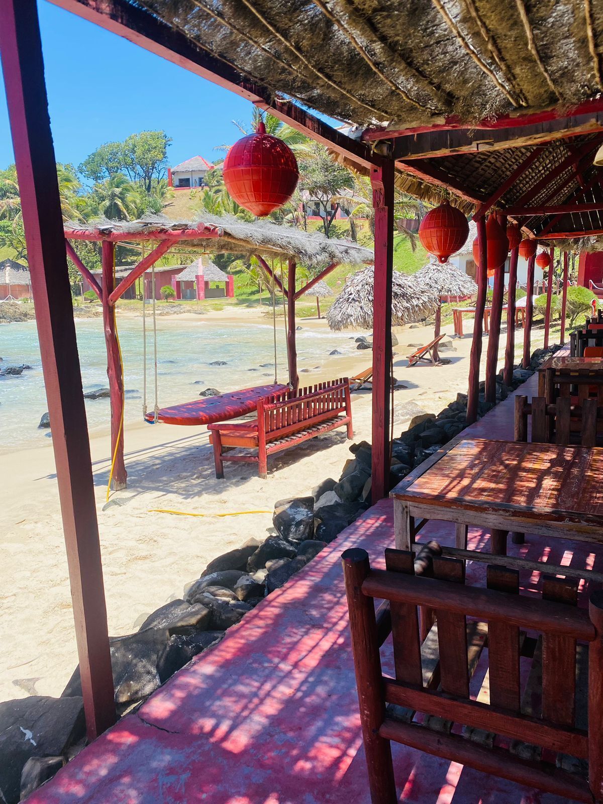 Un restaurante frente al mar con mesas y bancos de madera, techo de paja y faroles rojos.