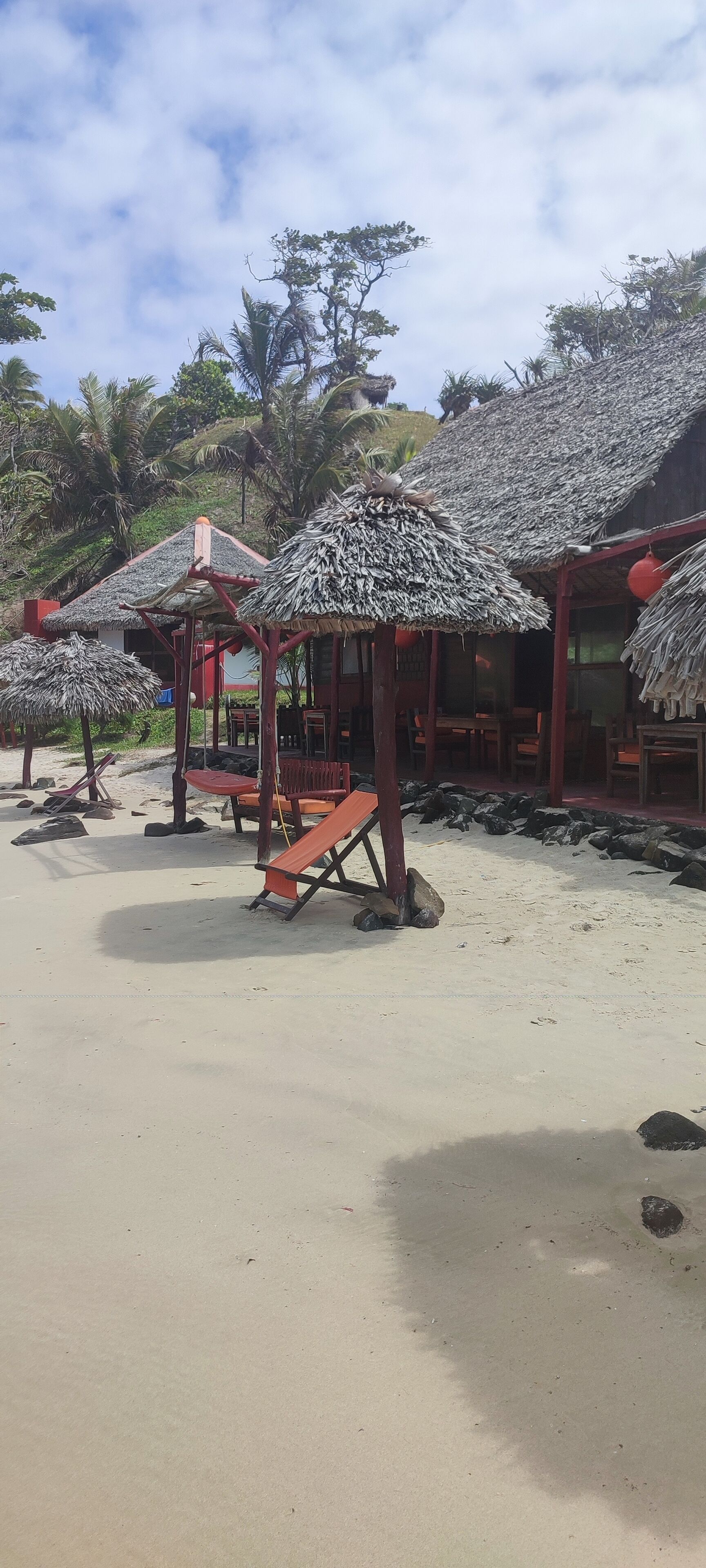Zona de playa con sombrillas de paja, mesas y sillas de madera.