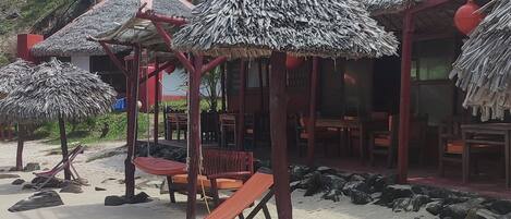 Zona de playa con sombrillas de paja, mesas y sillas de madera.