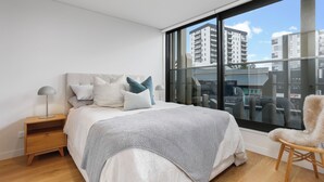 Un dormitorio moderno con un ventanal grande, una cama con manta gris y blanca, una mesita de noche de madera y una silla.