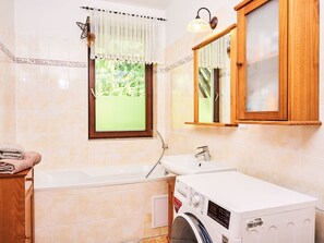Bathroom features a large bathtub and wall-mounted washing machine, with a window and storage cabinet.
