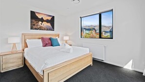 A bedroom with a wooden bed, two bedside tables, a large window with a view, and a painting on the wall. - Views on Farrant Summ (Wānaka)