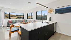 A modern kitchen with a large island, a dining area with a sofa, and large windows offering a view of the countryside. - Views on Farrant Summ (Wānaka)
