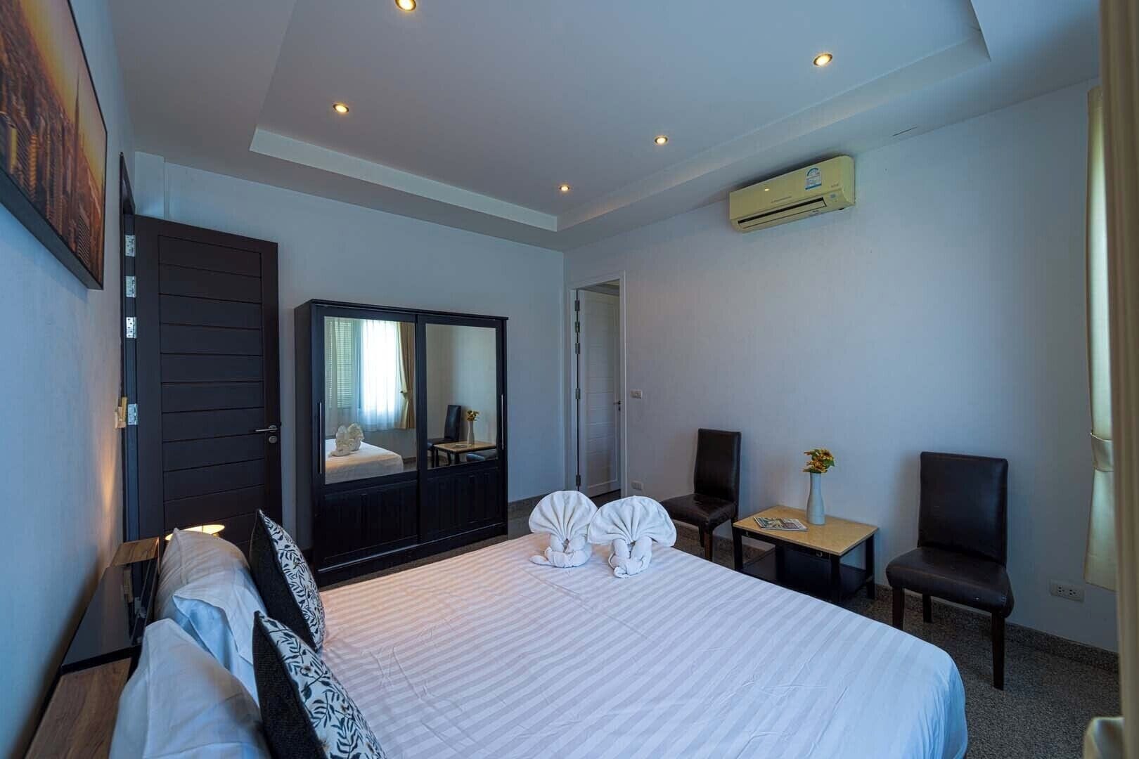 A hotel room with a large bed, a wardrobe, a desk, a chair, and an air conditioning unit.