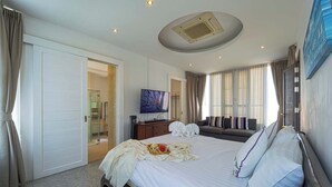 A hotel room with a large bed, a TV, a bathroom with a glass door, and a view of the city. - Luxury 4 Bedroom Pool Villa - PV8 (Hua Hin)