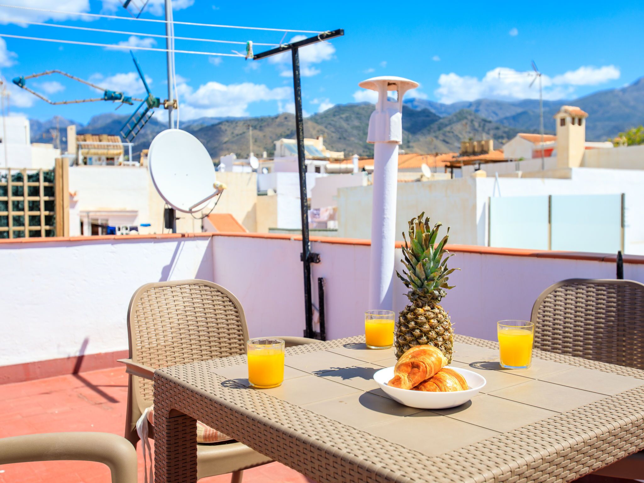 Terraço no telhado com móveis de vime, uma mesa com croissants e suco de laranja, uma abacaxi e vista para a cidade e montanhas.