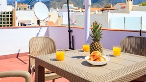 A rooftop terrace with wicker furniture, a table with croissants and orange juice, a pineapple, and a view of the cityscape and mountains. - Apartment Parra by Interhome (Nerja)