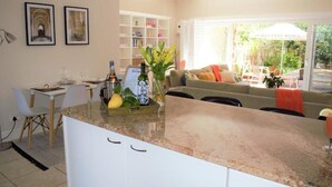 Una cocina moderna con encimera de granito, un comedor con mesa y sillas blancas, un sofá con cojines naranjas y una estantería.