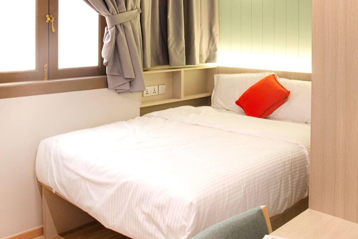 A neatly made bed with white bedding and a red pillow, a wooden nightstand with a lamp, and a window with curtains.