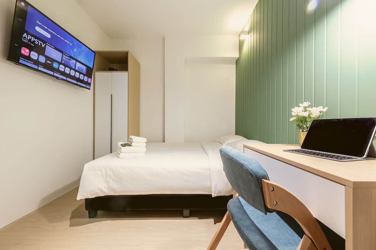 A hotel room with a bed, a desk with a laptop, a TV mounted on the wall, and a green wall panel.