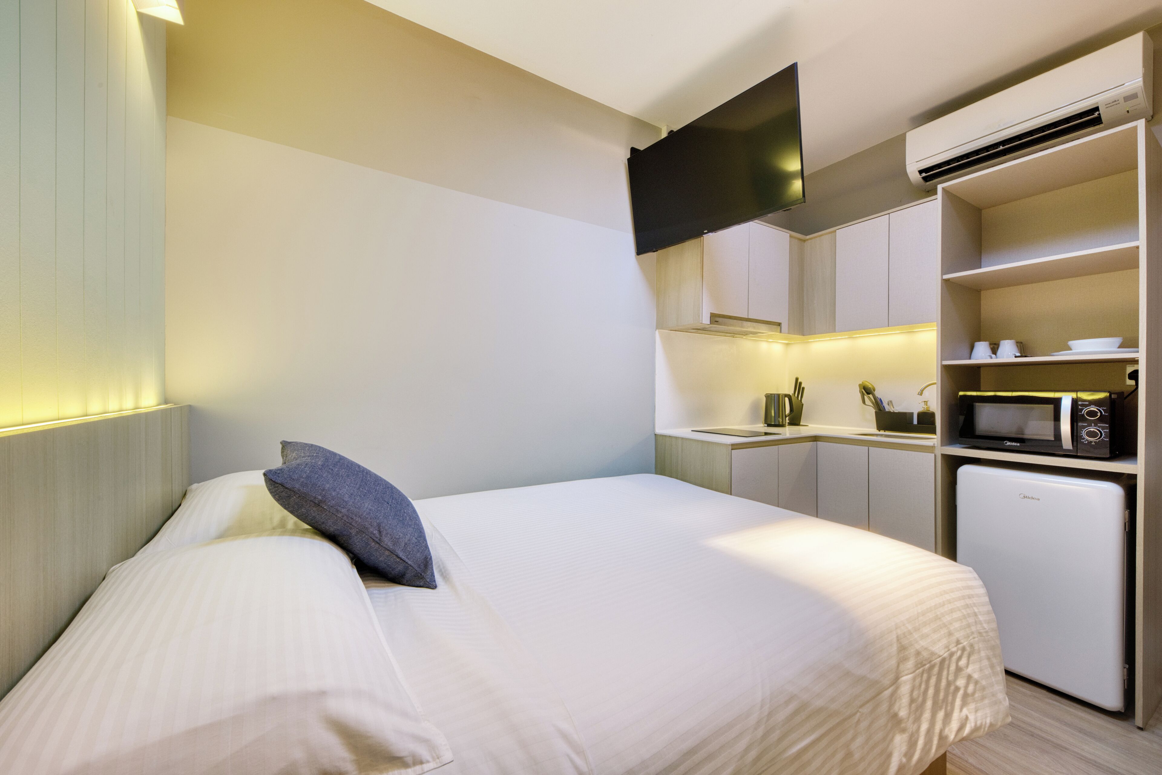 Une chambre d’hôtel comprenant un lit, un téléviseur à écran plat fixé au-dessus, une kitchenette équipée d’un four à micro-ondes et d’un réfrigérateur, ainsi qu’un petit placard contenant des ustensiles de cuisine.