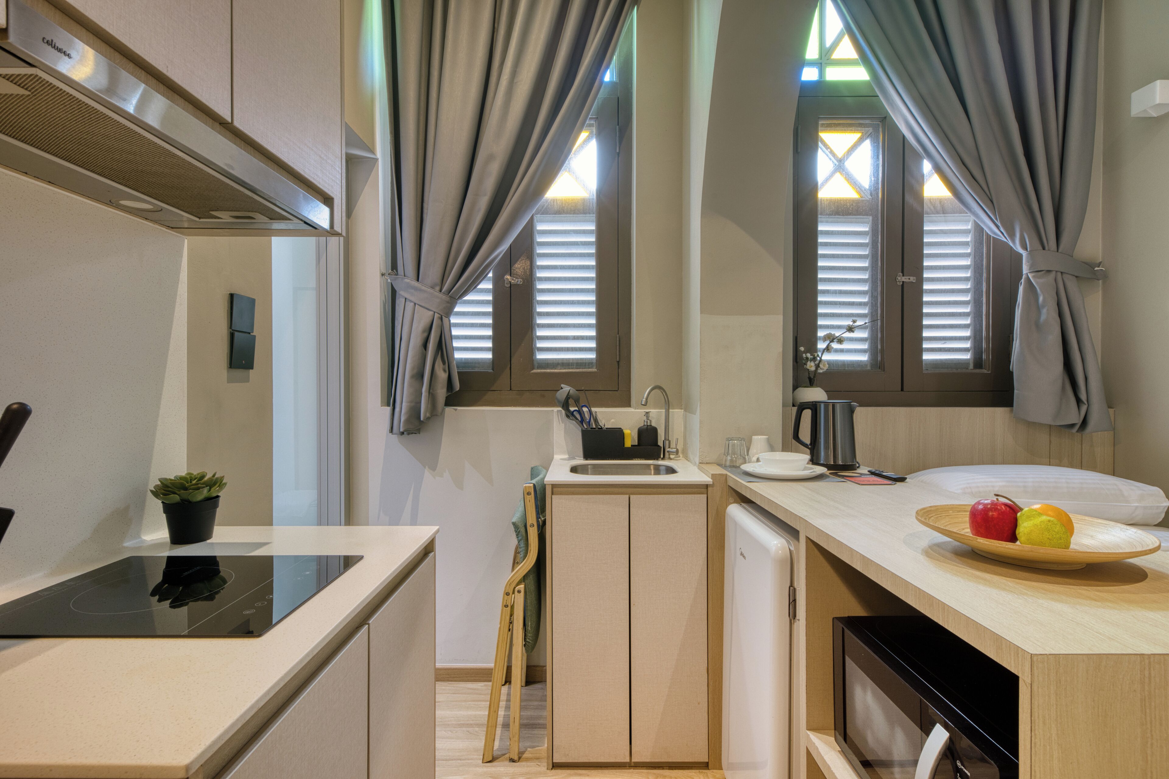 Cocina compacta con estufa integrada, pileta y heladera pequeña. Hay dos ventanas con persianas, una planta en maceta y un cuenco con fruta en la encimera.