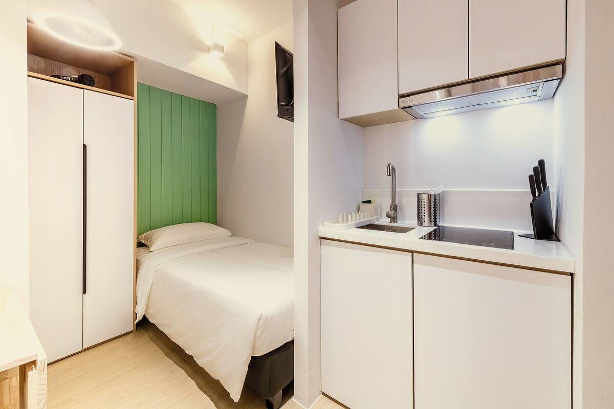 A compact hotel room with a bed, a kitchenette, and a green accent wall.