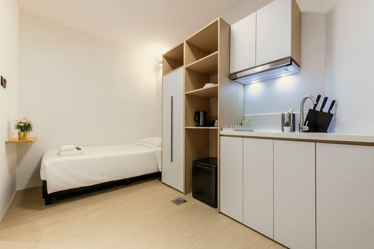 A compact room with a bed, kitchenette, and a small shelf with a plant.