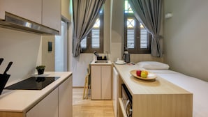 Une petite cuisine équipée avec une cuisinière, un évier et un petit plan de travail. On y trouve un lit avec une tête de lit en bois et une table de chevet sur laquelle est posé un panier de fruits.