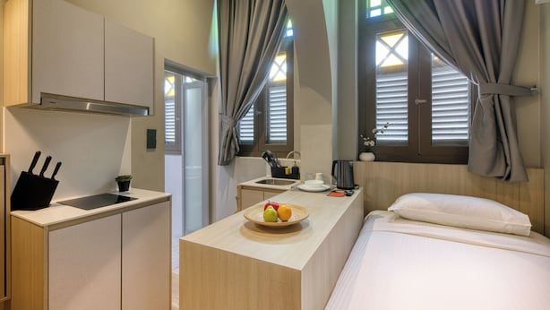 Une chambre d’hôtel compacte comprenant un coin cuisine, un lit avec du linge de maison blanc, une petite table à manger avec des fruits, et une vue sur l’extérieur par une fenêtre équipée de stores.