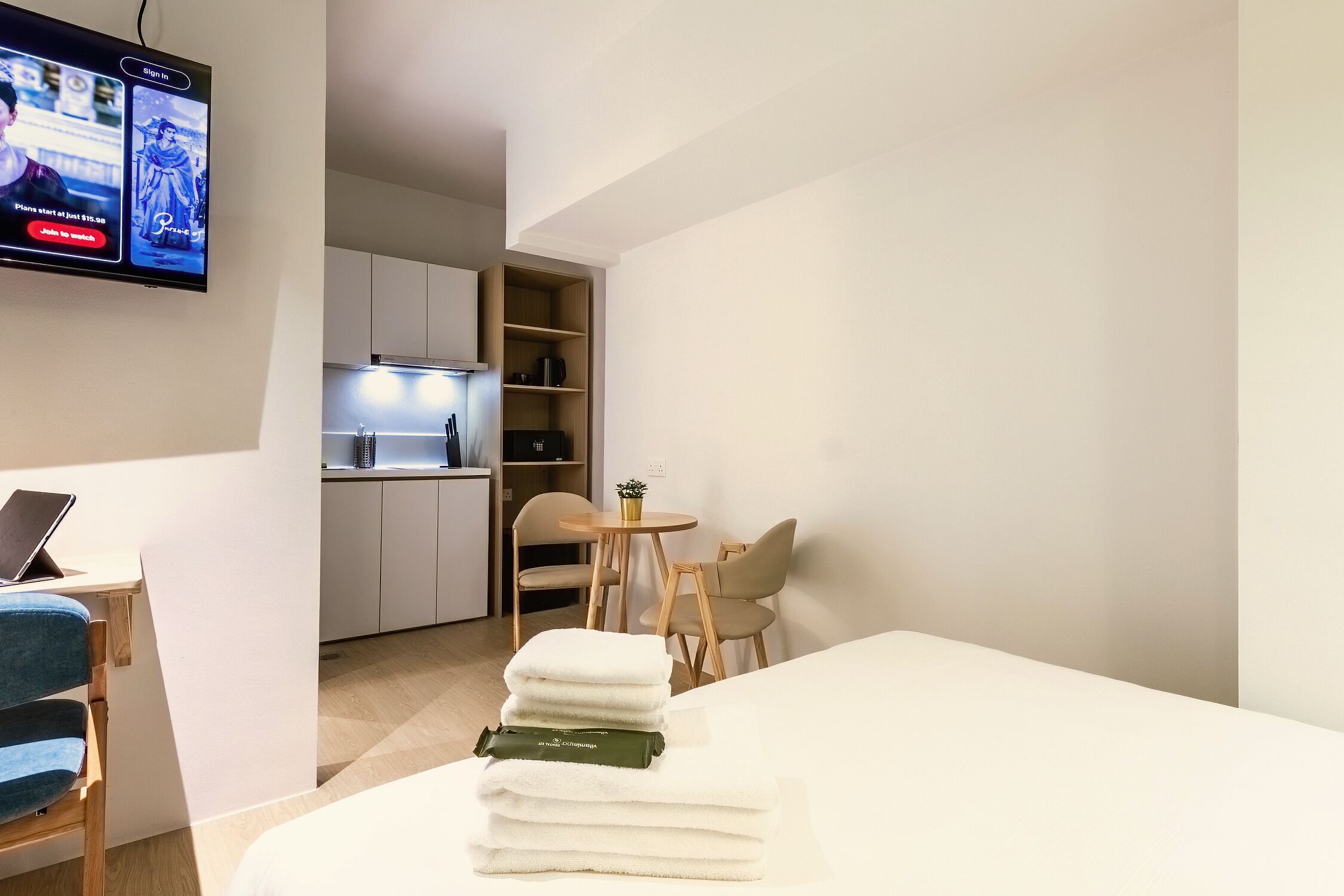 A hotel room with a bed, a desk with a laptop, a TV, a small kitchen area, and a dining table with chairs.