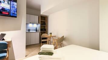 A hotel room with a bed, a desk with a laptop, a TV, a small kitchen area, and a dining table with chairs.