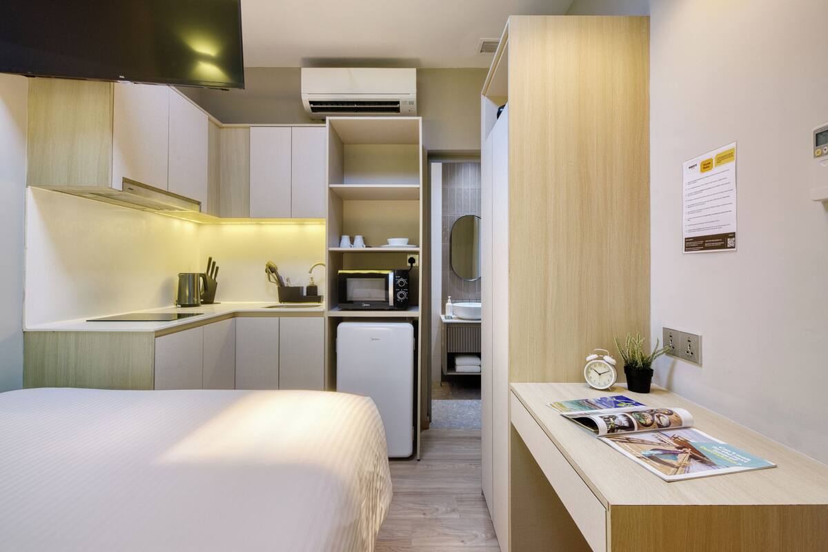 A compact hotel room with a bed, kitchenette, and a small table with a magazine.