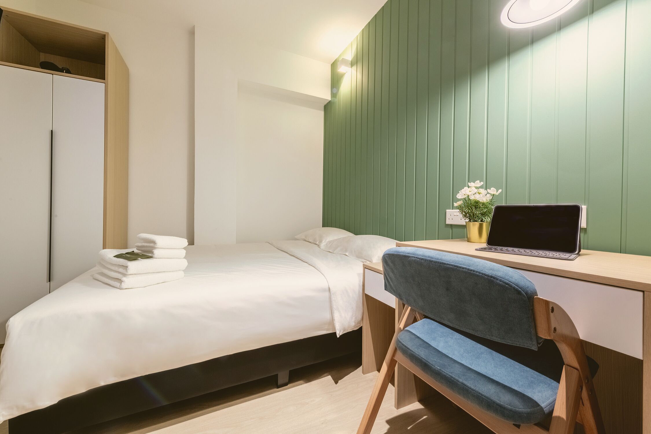 A bedroom with a bed, a desk with a laptop, a chair, and a wardrobe.