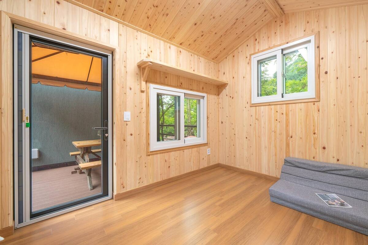 A wooden cabin interior with a bench, two windows, and a door leading to an outdoor area.