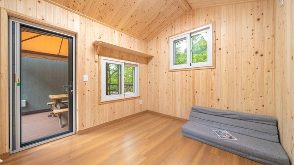 A wooden cabin interior with a bench, two windows, and a door leading to an outdoor area.