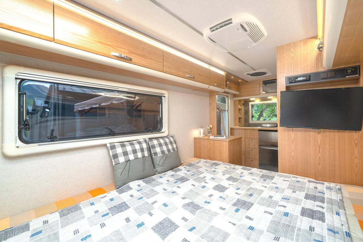 A compact RV interior with a bed, kitchenette, and a TV mounted on the wall.