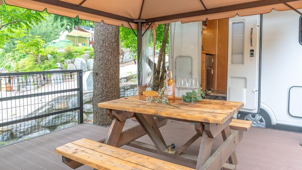 Mesa de picnic de madera con bancos, una botella de vino y vasos en un área exterior cubierta con vistas a una caravana blanca y follaje al fondo.