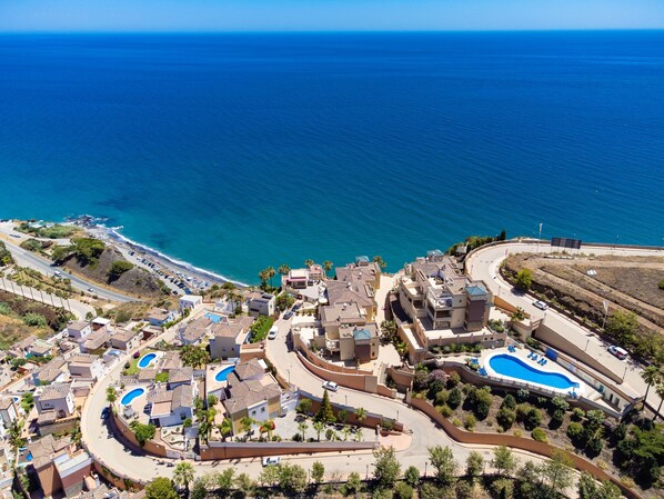 An aerial view of a coastal residential area with multiple swimming pools, a curved road, and a beach.