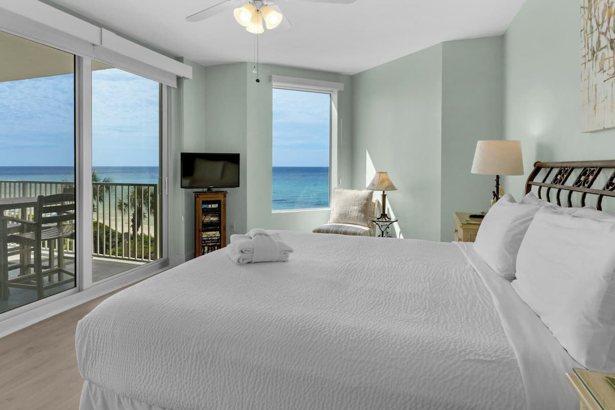 A bedroom with a large bed, a TV, a nightstand, and a view of the ocean.