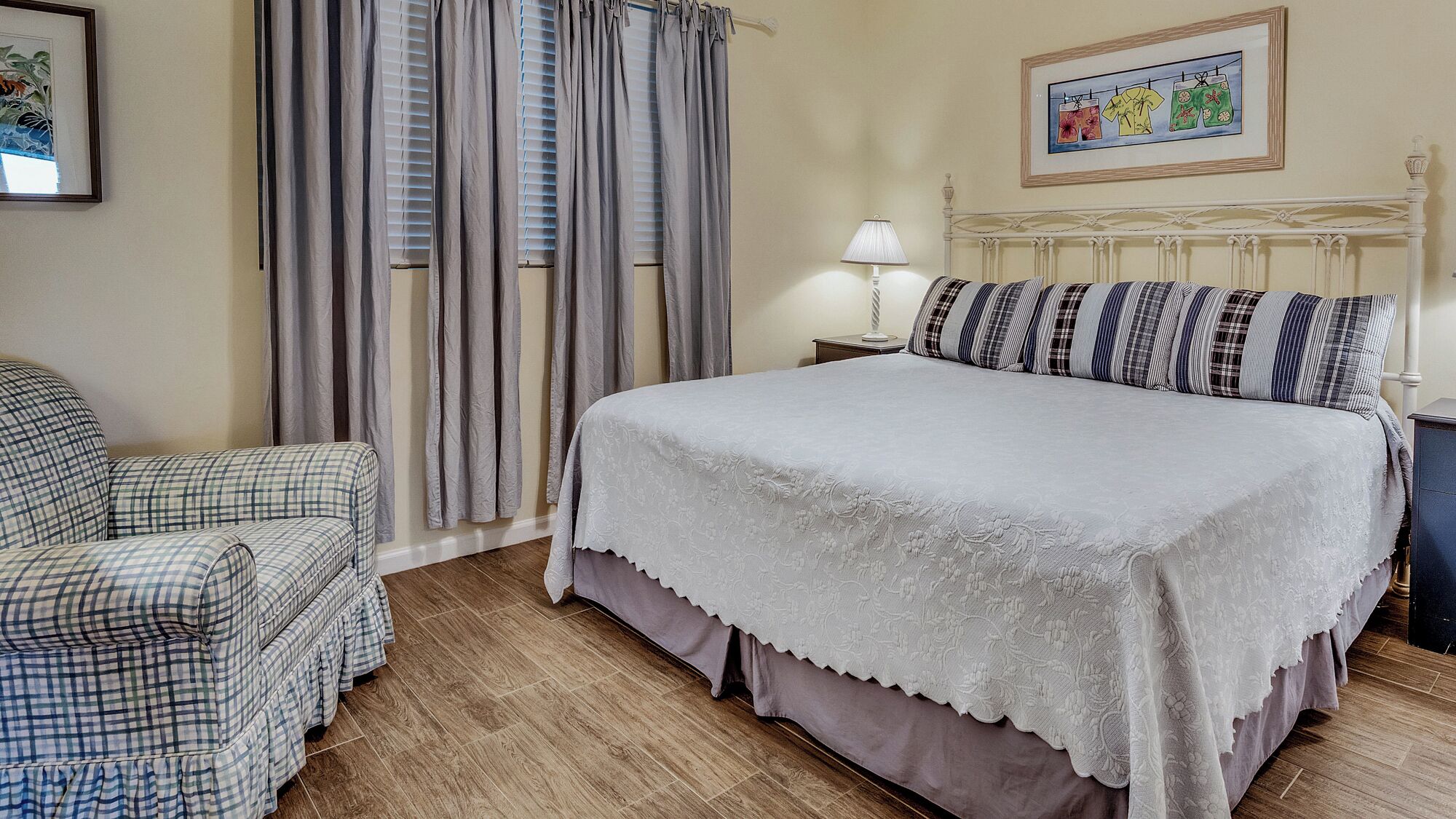 A hotel room with a large bed, a plaid armchair, a bedside table with a lamp, and a painting on the wall.