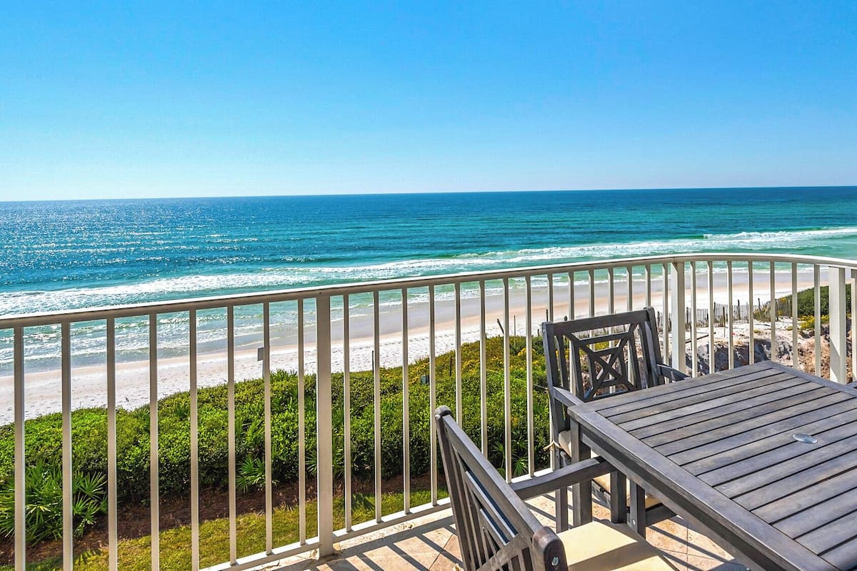 A balcony with a view of the ocean, a wooden table, and chairs.