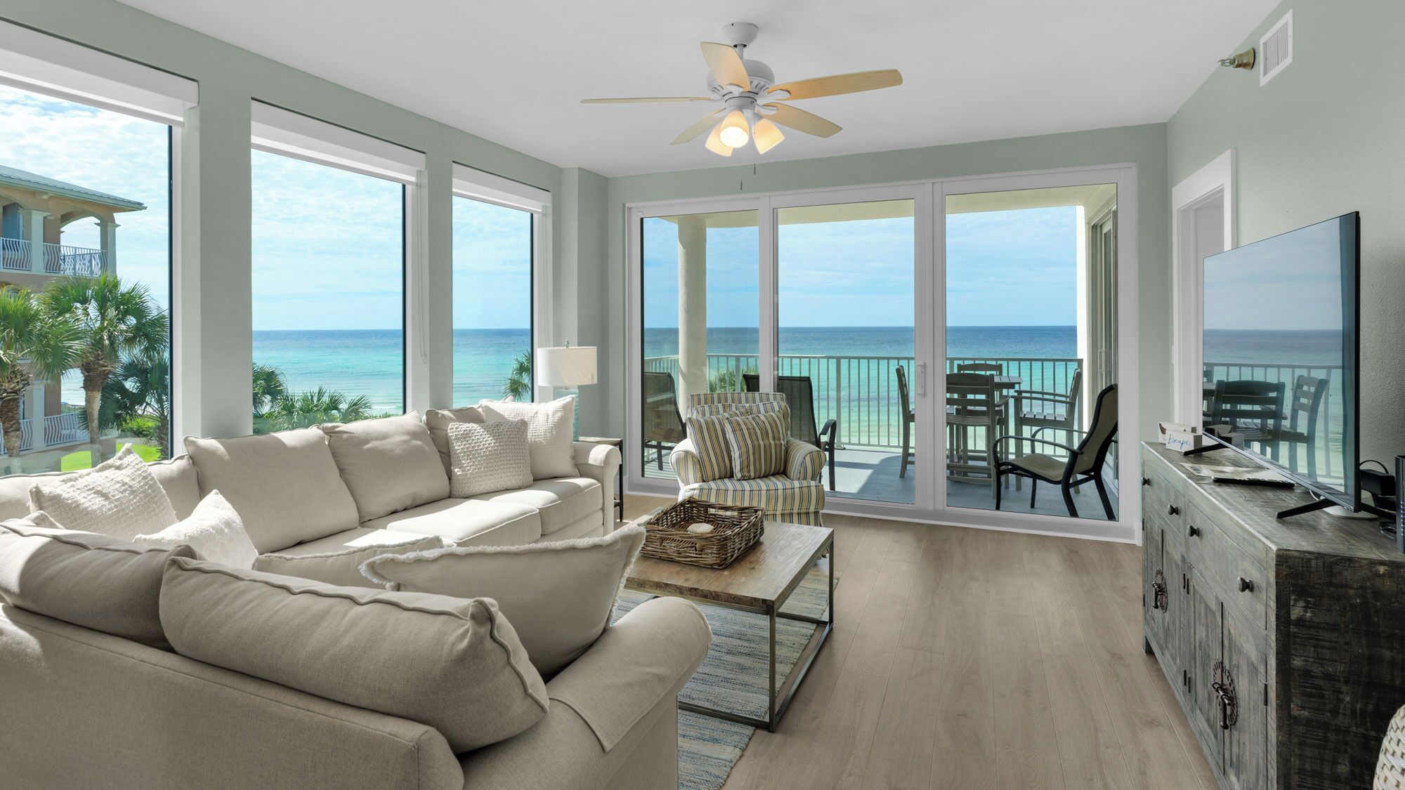A spacious living room with a large sofa, a coffee table, and a TV on a wooden cabinet. The room has large windows offering a view of the ocean and a balcony with outdoor furniture.