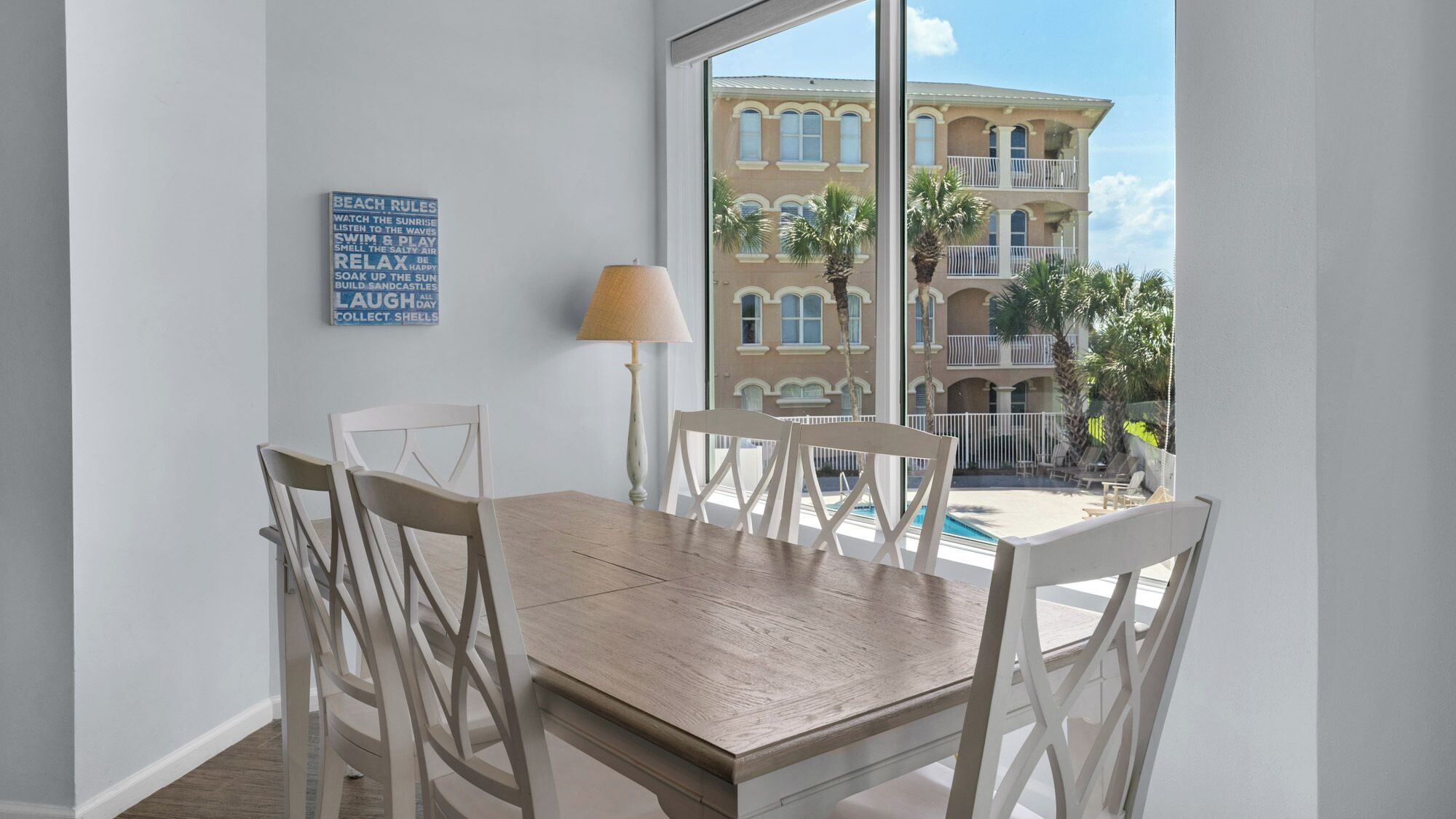 A dining area with a wooden table and white chairs, a lamp, and a view of a building with balconies and palm trees.