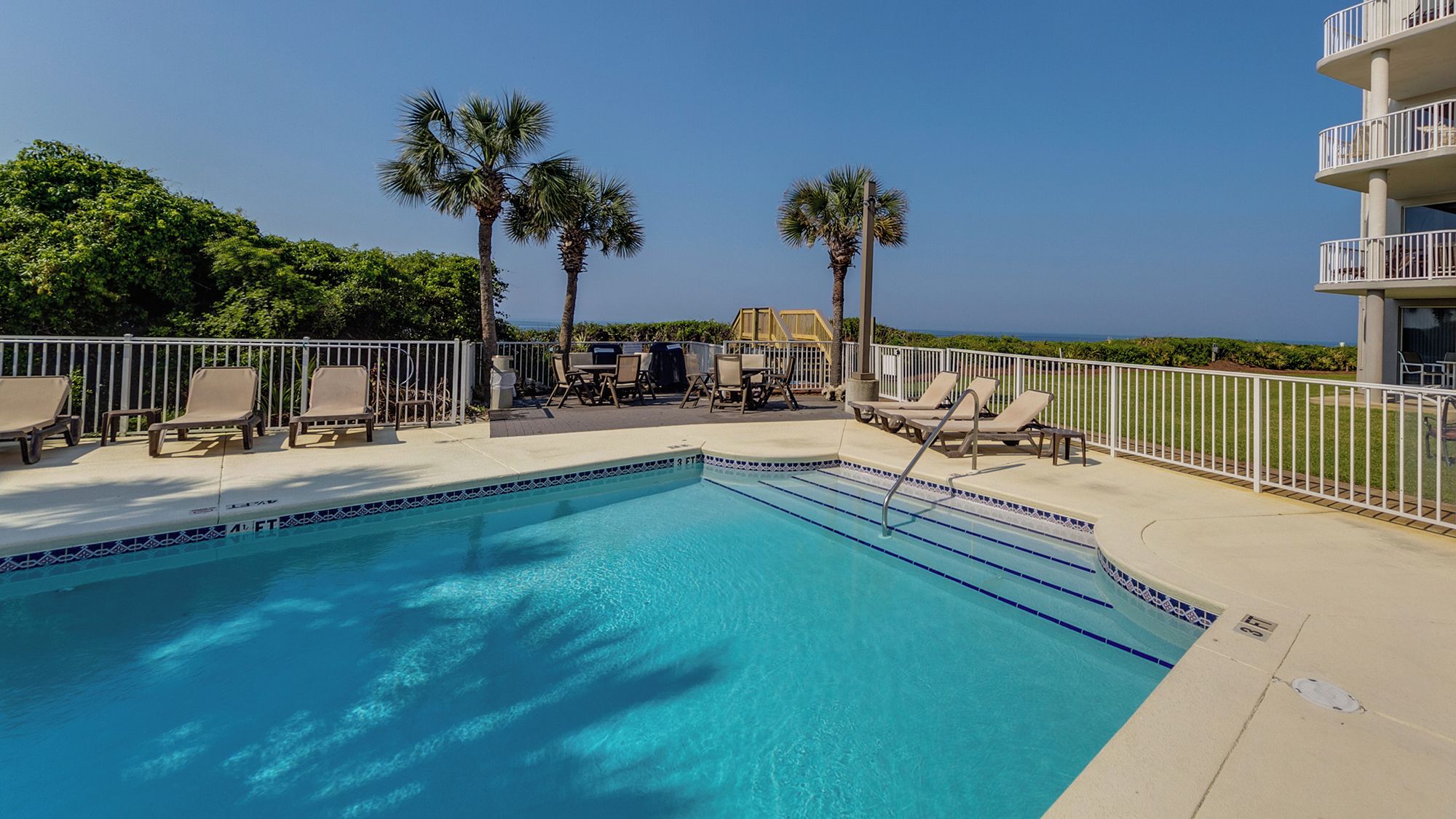 A swimming pool with lounge chairs and palm trees, surrounded by a white fence.