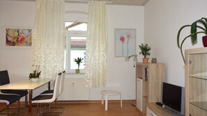 Una habitación con una mesa de comedor, sillas, una mesita blanca, un mueble con televisor, una planta en maceta y un ventanal con cortinas.