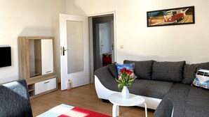 Una sala de estar con un sofá, una mesita blanca, un televisor y una puerta que da a otra habitación.