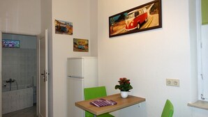 Una kitchenette pequeña con un refrigerador, una mesa con una planta en maceta y una pared con un cuadro enmarcado.