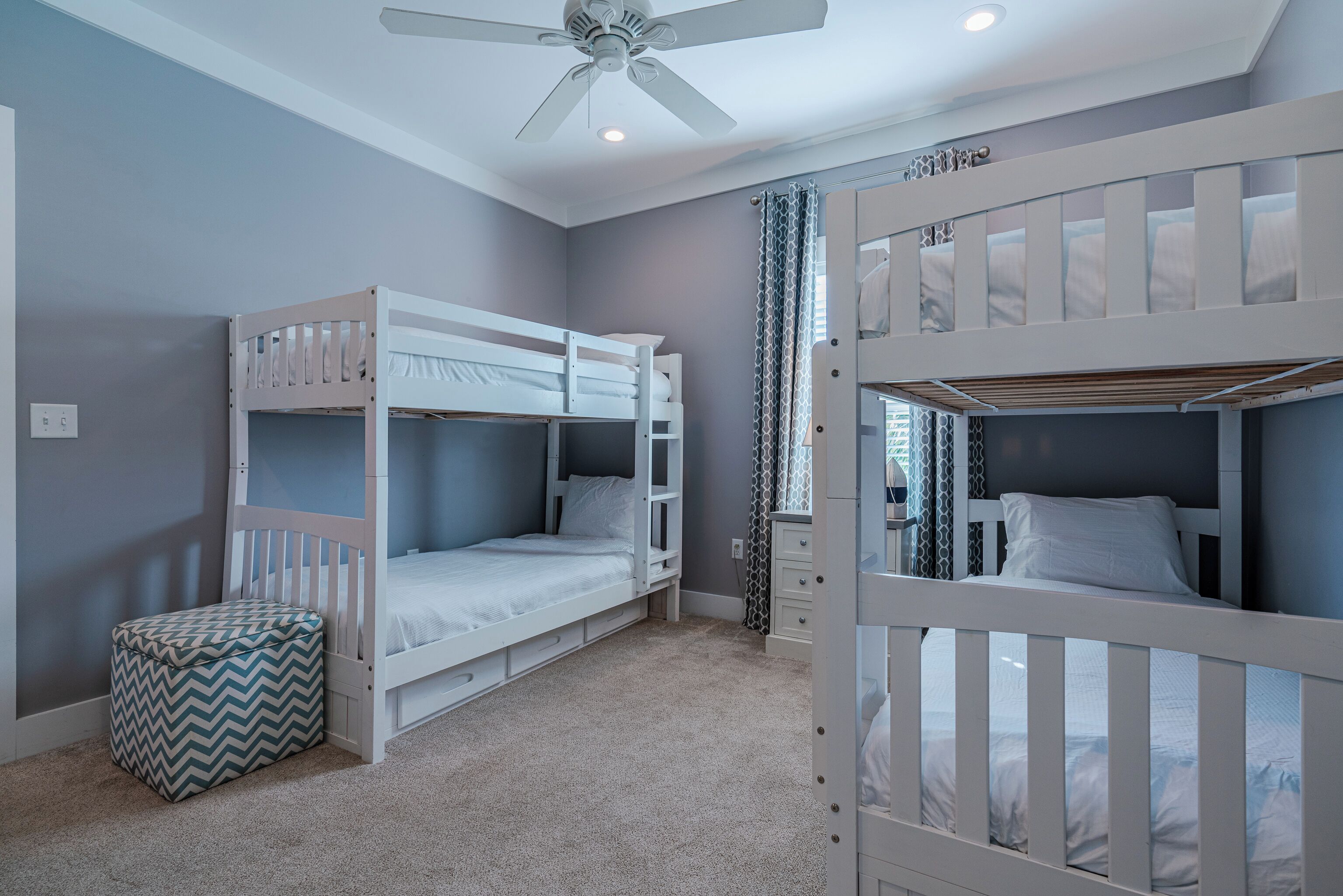 Une chambre d’enfant avec deux lits superposés, un ventilateur de plafond et un petit pouf.