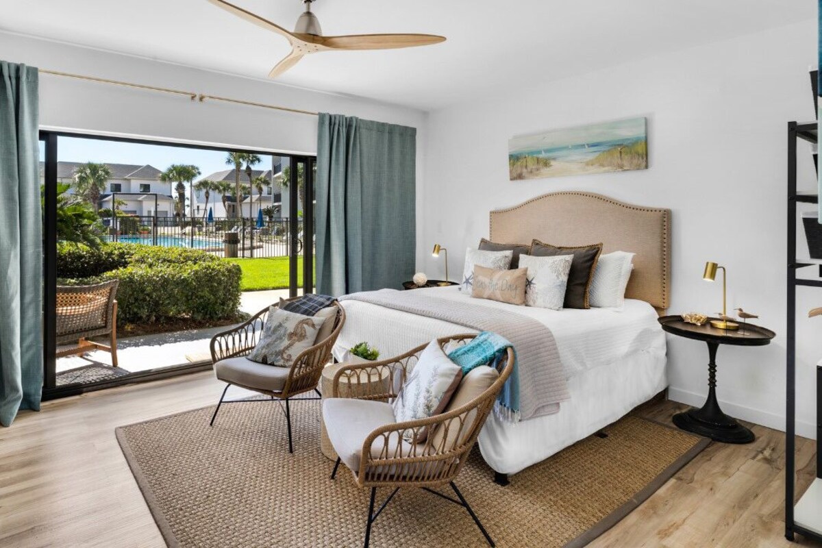 Une chambre d’hôtel moderne avec un grand lit, un balcon donnant sur une piscine et des immeubles, des meubles en osier et un ventilateur de plafond.