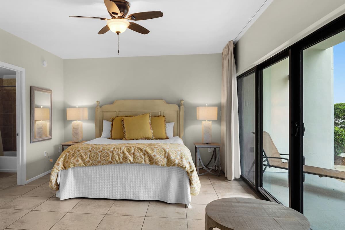 A bedroom with a bed, bedside lamps, a ceiling fan, and a view through a large window.