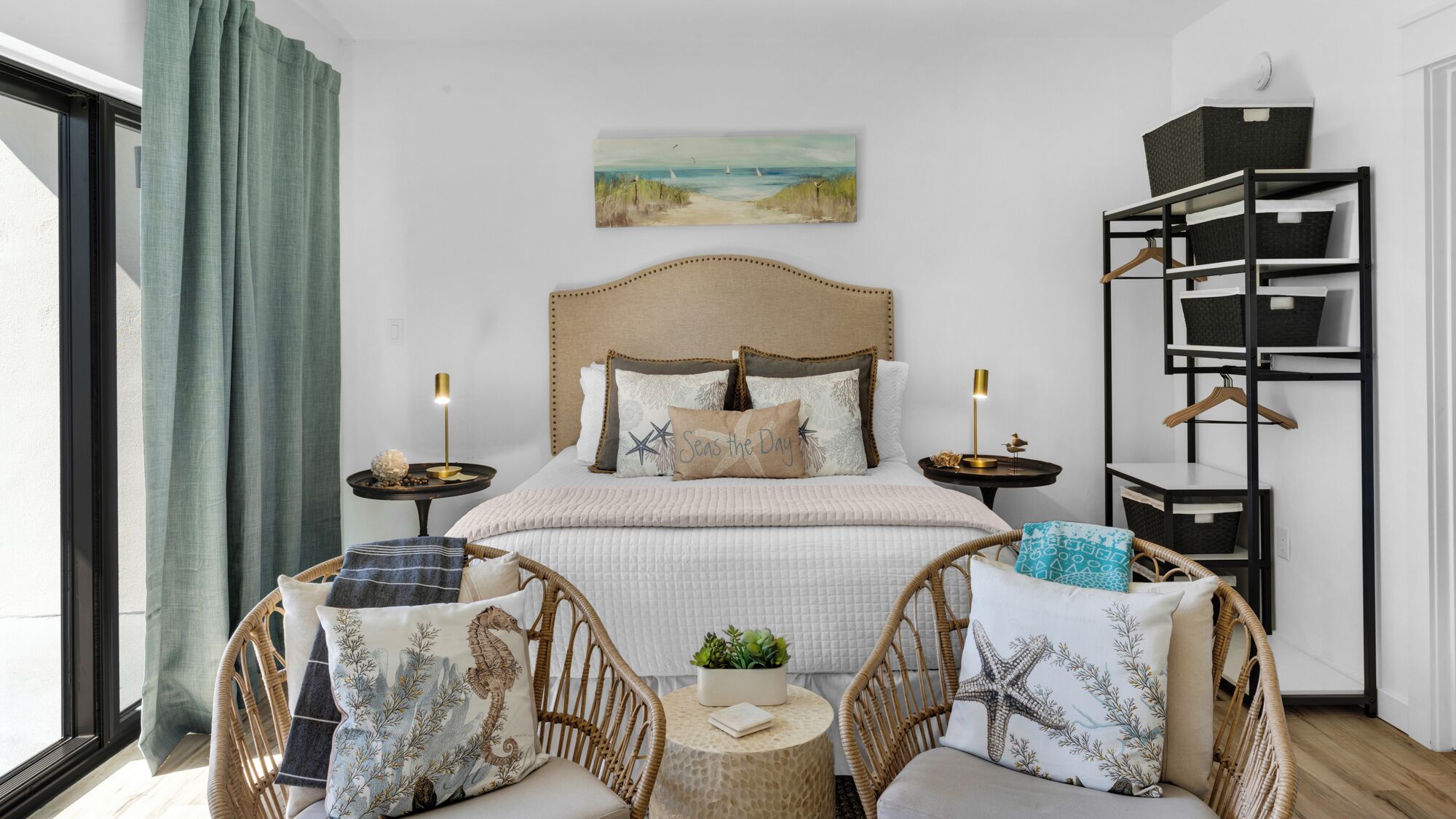 A bedroom with a bed, two nightstands, a wall-mounted shelf, a wicker chair, and a view of the outdoors.