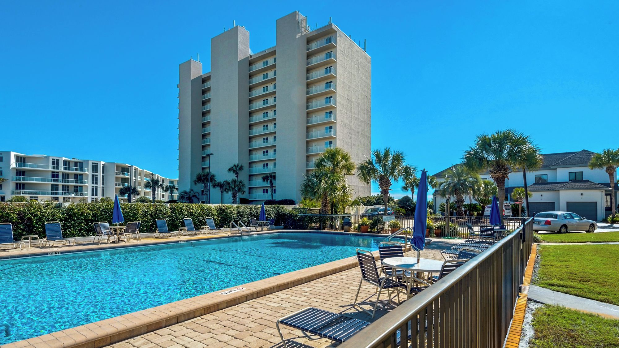 A swimming pool with lounge chairs and umbrellas, surrounded by a fence, with a tall building in the background.