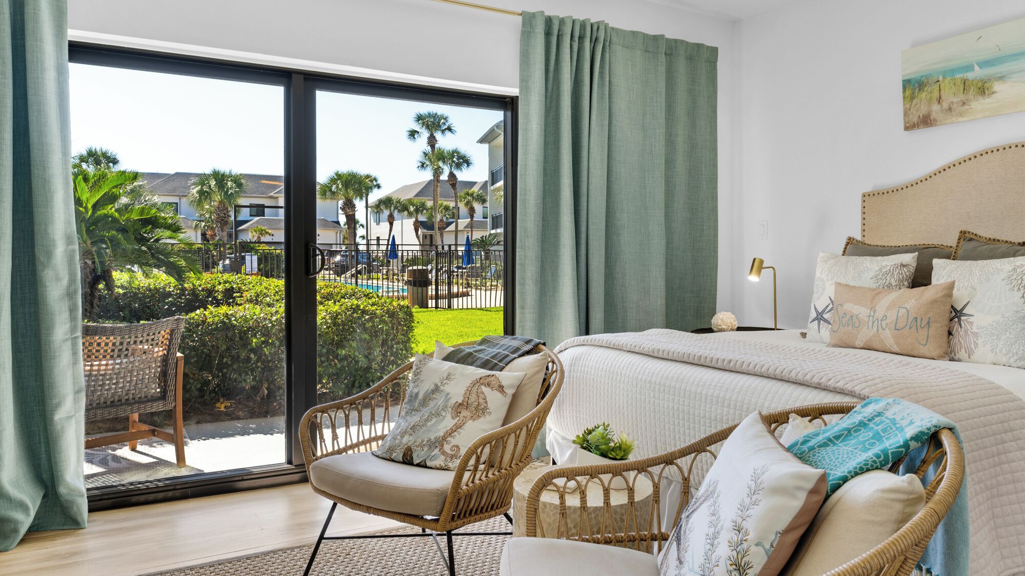 A bedroom with a large bed, wicker chairs, a view of a pool and palm trees, and a painting on the wall.