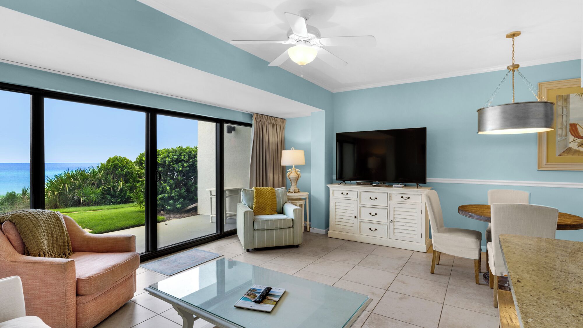 A hotel room with a large sliding glass door leading to a balcony with a view of the ocean. There is a sofa, a coffee table, a TV, and a dining area with a table and chairs.