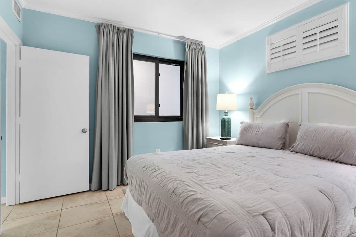 A bedroom with a bed, curtains, a nightstand, a lamp, and a window.
