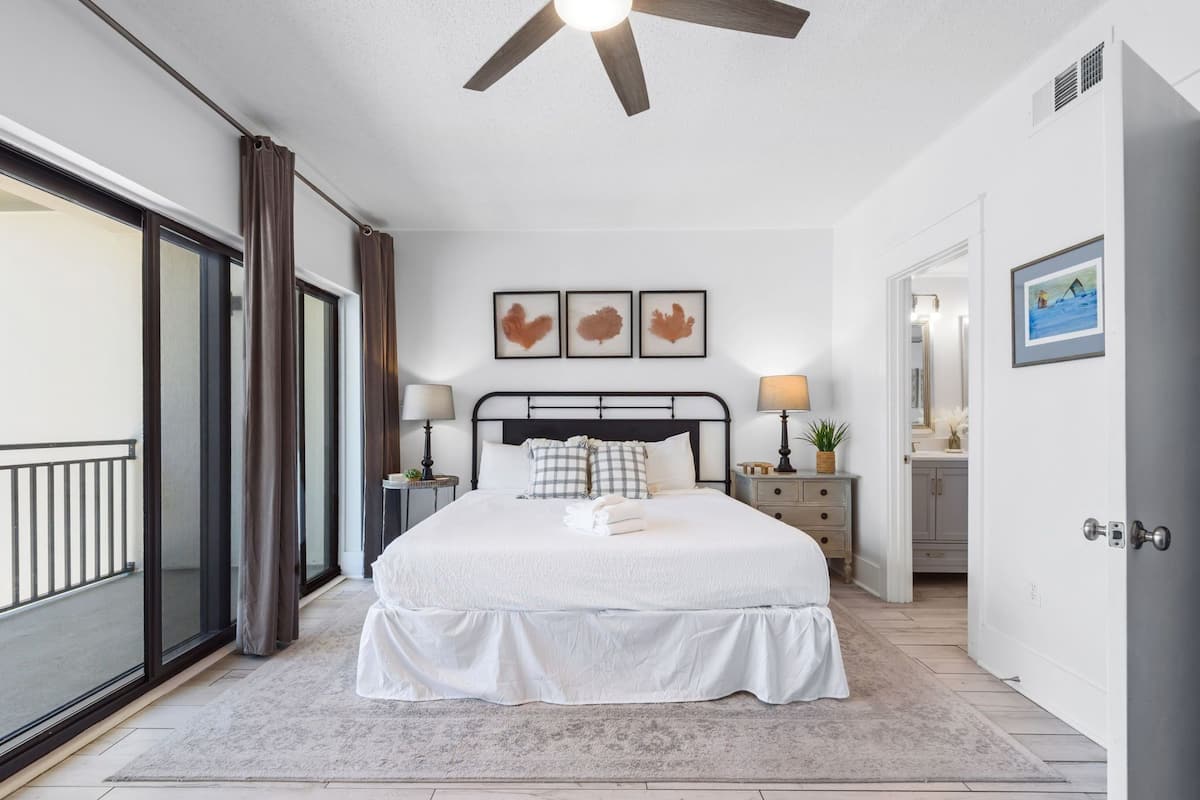 A bedroom with a bed, a ceiling fan, a large window, and a door leading to a bathroom.