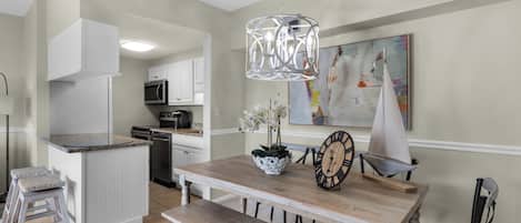 A kitchen with white cabinets, a microwave, and a coffee maker. There is a dining table with a bench, a potted orchid, and a clock.